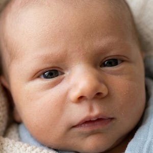 May include: A close-up portrait of a newborn baby with fair skin and dark eyes. The baby is wrapped in a soft, cream-coloured blanket and wearing a light blue garment. The baby's expression is serious.