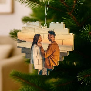 May include: Heart-shaped Christmas ornament made of interlocking blocks, featuring a photograph of a couple. The ornament hangs from a Christmas tree branch. The photo shows a couple at the beach during sunset.