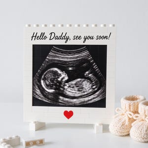 May include: A white building block frame displaying a black and white ultrasound image of a fetus. The frame features the text Hello Daddy, see you soon! in black script and a small red heart icon. A pair of cream-colored knit baby booties sits nearby.
