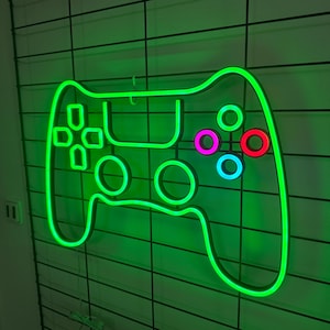 May include: A green neon light shaped like a video game controller with colourful buttons. The controller is mounted on a white wall with a grid pattern.