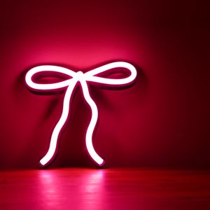 Peut inclure: Un signe néon blanc en forme de nœud, qui brille en rose sur un fond rouge.