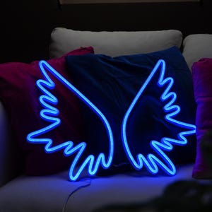 May include: A blue neon light shaped like angel wings. The wings are displayed against a backdrop of pillows in shades of pink, blue, and white. The neon light is plugged in and ready to illuminate.