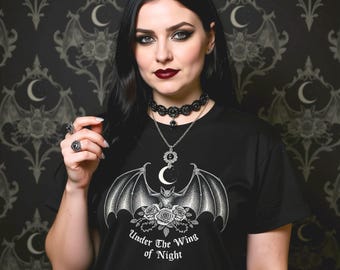 Op de afbeelding: Zwart T-shirt met een vleermuis- en rozenafbeelding en de tekst "Under The Wing of Night". De afbeelding toont ook een zwarte choker, een hangerketting met een maan en een ring met een zwarte steen. De achtergrond heeft een donker patroon.