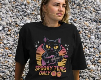May include: Black t-shirt featuring a cartoon cat playing a pink guitar against a sunset backdrop. The cat wears a leather jacket. Text on the shirt reads "Spooky Tunes Only" with two smiling ghost emojis. The shirt is worn by a person.