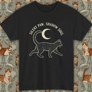 Könnte beinhalten: Schwarzes T-Shirt mit einer weißen Illustration einer Katze und einem Mond. Der Text "SILENT PAW, SHADOW SOUL" ist darüber angeordnet. Das T-Shirt besteht aus einem weichen Material.