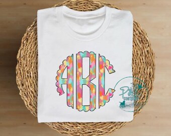May include: White t-shirt with a colorful monogram design. The monogram is in a circle with a scalloped edge and features the letters "ABC" in a colorful abstract pattern.