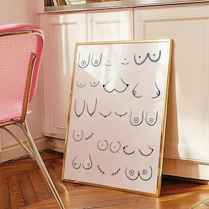 以下が含まれることがあります： さまざまな形をした乳房の黒い線画が描かれた、金色の縁取りのある額入りのプリント。プリントは木の床に表示されています。フレームはピンクの椅子の近くにあります。