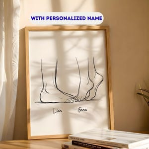 May include: A framed art print featuring a minimalist line drawing of two pairs of feet, with the names "Liam" and "Emma" below. The text "WITH PERSONALIZED NAME" is at the top. The artwork is in a light-colored wooden frame.