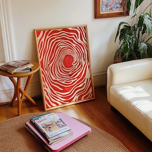 May include: A framed abstract art print featuring a red and gold concentric pattern. The artwork is displayed in a gold frame and leans against a white wall. The print's design resembles a target or a ripple effect.
