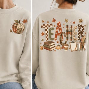May include: A beige crewneck sweatshirt featuring a small patchwork apple graphic on the front left chest. The back displays the word TEACHER in patterned letters, surrounded by autumn leaves, a stack of books, a pumpkin, and a pencil jar.
