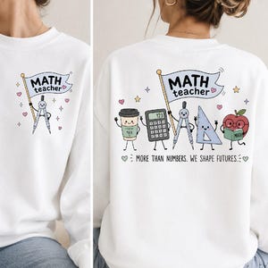 May include: A white crewneck sweatshirt featuring a small graphic on the front left chest of a compass holding a flag that reads MATH teacher. The back displays a larger graphic of a coffee cup, calculator, compass, ruler, and apple with the text MATH teacher and MORE THAN NUMBERS. WE SHAPE FUTURES.
