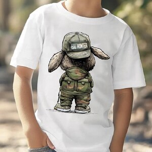 Puede incluir: Camiseta blanca con una ilustraci&oacute;n de dibujos animados de un conejito con un atuendo de camuflaje y una gorra que dice "EGG HUNTER". El conejito se representa de espaldas, con sus orejas y detalles del atuendo claramente visibles. El dise&ntilde;o est&aacute; centrado en la camiseta.