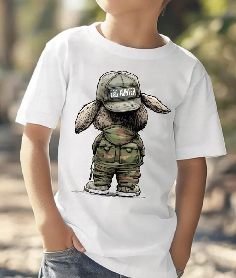 Puede incluir: Camiseta blanca con una ilustraci&oacute;n de dibujos animados de un conejito con un atuendo de camuflaje y una gorra que dice "EGG HUNTER". El conejito se representa de espaldas, con sus orejas y detalles del atuendo claramente visibles. El dise&ntilde;o est&aacute; centrado en la camiseta.