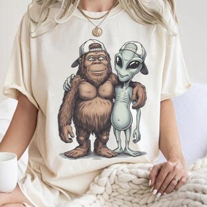 May include: Cream-colored t-shirt featuring a cartoon graphic of a Bigfoot and an alien with their arms around each other, both wearing backward baseball caps. The Bigfoot is brown and furry, while the alien is gray. The shirt has a relaxed fit.
