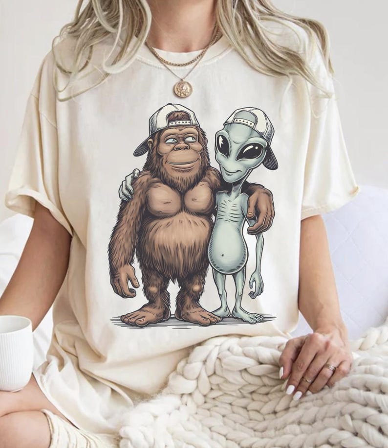 May include: Cream-colored t-shirt featuring a cartoon graphic of a Bigfoot and an alien with their arms around each other, both wearing backward baseball caps. The Bigfoot is brown and furry, while the alien is gray. The shirt has a relaxed fit.