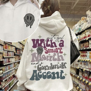 Puede incluir: Sudadera con capucha blanca con un estampado gráfico en la espalda que dice "Just a girl with a smart mouth and a cornbread accent". El gráfico está en un estilo retro con una cara sonriente que gotea.