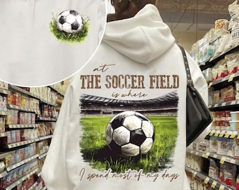 May include: White hoodie with a brown graphic of a soccer field and a soccer ball. The text on the hoodie reads "At the soccer field is where I spend most of my days."