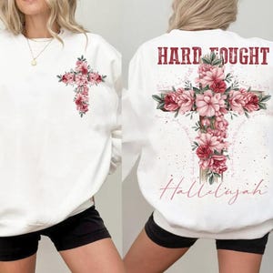 Może przedstawiać: Biała bluza z kwiatowym wzorem krzyża z przodu i napisem "Hard Fought Hallelujah" z tyłu.