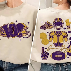 Op de afbeelding: Een wit sweatshirt met een paars en gouden voetbalontwerp op de voorkant. Het ontwerp bevat een voetbal, een helm, een schoen en een megafoon. De achterkant van het sweatshirt heeft een paars en gouden ontwerp met een voetbalhelm, een voetbal, een megafoon, een shirt en pompoenen. De tekst op de achterkant van het sweatshirt zegt "Game Day" en "Touch Down Season".