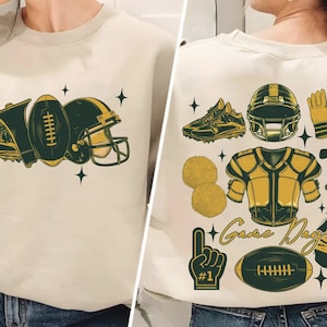May include: A white sweatshirt with a green and gold football design on the front. The design includes a football, a helmet, a shoe, and a whistle. The back of the sweatshirt has a green and gold design with a football, a helmet, a shoe, a cheerleader's pom-pom, a football jersey, a megaphone, and a hand with a number one finger raised. The text "Game Day" and "Touch Down Season" are also included in the design.