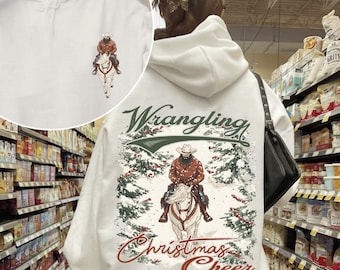 May include: White hoodie with a graphic of a cowboy riding a horse through a snowy forest with the text "Wrangling Christmas Cheer" on the back.