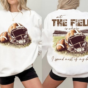 May include: White sweatshirt with a brown football helmet and football graphic on the front. The back of the sweatshirt has the text "At the field is where I spend most of my days".
