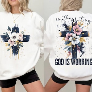 May include: White sweatshirt with a blue and black cross with floral accents. The text "in the waiting GOD IS WORKING" is printed on the back of the sweatshirt.