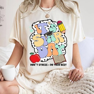 May include: A white t-shirt with a colorful graphic that says "It's Star Day" with a graduation cap, pencil, apple, and open book. The text below the graphic says "Don't Stress - Do Your Best".