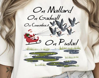 May include: White t-shirt with a Christmas-themed design featuring Santa Claus in a sleigh pulled by a flock of ducks. The text reads "On Mallard On Gadwall On Canvasback On Pintail".