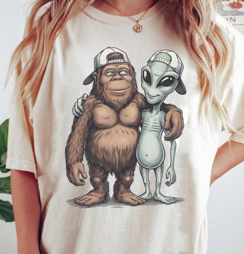 May include: Cream-colored t-shirt featuring a cartoon illustration of a Bigfoot character and an alien, both wearing white baseball caps. The Bigfoot has brown fur, and the alien is gray. They are embracing each other.