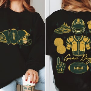 May include: Black sweatshirt with a green and gold football theme design. The design includes a football, helmet, shoe, and other football-related imagery. The text "Game Day" is included in the design.