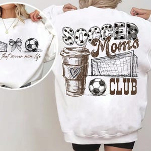 Może przedstawiać: Biała bluza z motywem piłkarskim. Prz&oacute;d przedstawia piłkę nożną, kokardę i bramkę. Tył przedstawia "Soccer Moms Club" z filiżanką kawy, piłką nożną i grafiką bramki. Projekt jest w kolorze czarnym i brązowym.