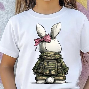 Puede incluir: Camiseta blanca con una ilustraci&oacute;n de conejo de dibujos animados. El conejo lleva una chaqueta y mochila de camuflaje, y un lazo rosa. El dise&ntilde;o es un gr&aacute;fico caprichoso y juguet&oacute;n, adecuado para ni&ntilde;os.