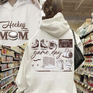 Puede incluir: Sudadera con capucha blanca con texto marr&oacute;n y gr&aacute;ficos de hockey. La parte delantera presenta "Hockey MOM" con un palo y un disco de hockey. La parte trasera muestra patines, un casco y las palabras "game day".