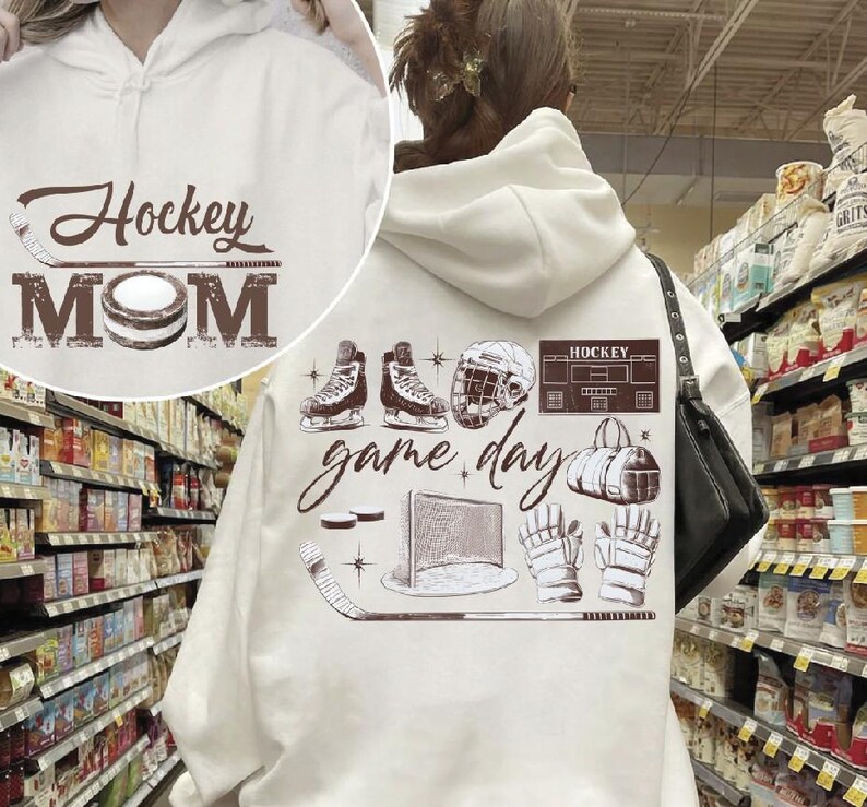 Puede incluir: Sudadera con capucha blanca con texto marr&oacute;n y gr&aacute;ficos de hockey. La parte delantera presenta "Hockey MOM" con un palo y un disco de hockey. La parte trasera muestra patines, un casco y las palabras "game day".