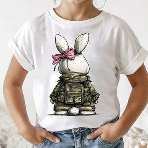 Puede incluir: Camiseta blanca con un conejito de dibujos animados que lleva una chaqueta de camuflaje y un lazo rosa. El conejito tiene orejas grandes y una cola esponjosa. El dise&ntilde;o es un gr&aacute;fico caprichoso y &uacute;nico.