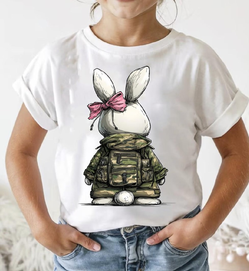 Puede incluir: Camiseta blanca con un conejito de dibujos animados que lleva una chaqueta de camuflaje y un lazo rosa. El conejito tiene orejas grandes y una cola esponjosa. El dise&ntilde;o es un gr&aacute;fico caprichoso y &uacute;nico.