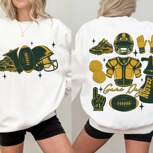May include: White sweatshirt with a green and gold football theme. The front of the sweatshirt has a football, helmet, and shoe. The back of the sweatshirt has a football helmet, football, megaphone, pom poms, and the text "Game Day Touch Down Season".