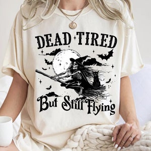 May include: A white t-shirt with a black graphic of a skeleton witch flying on a broomstick with a black cat. The text on the shirt reads "Dead Tired But Still Flying".