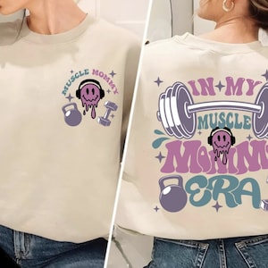 May include: A light beige sweatshirt with the text "Muscle Mommy Era" in pink, blue, and purple. The text is surrounded by dumbbells, a smiling face with headphones, and stars.