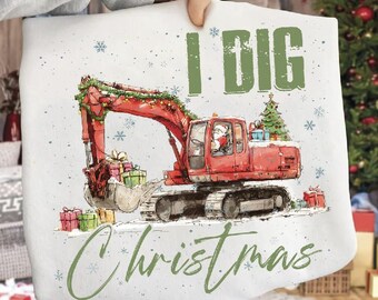 Puede incluir: Una sudadera blanca con una excavadora roja decorada con luces navideñas y un pequeño árbol de Navidad. Papá Noel está en el asiento del conductor. El texto "I DIG Christmas" está impreso en verde.
