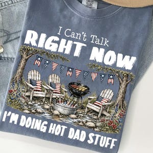 May include: A blue Comfort Colors t-shirt with the text "I Can't Talk Right Now I'm Doing Hot Dad Stuff." The graphic depicts a backyard scene with Adirondack chairs, a grill, and American flag decorations.