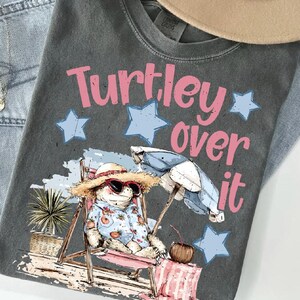 May include: Dark gray t-shirt with the phrase "Turtley over it" in pink lettering and blue stars. The graphic features a turtle wearing sunglasses and a straw hat, relaxing in a beach chair with an umbrella and a drink.