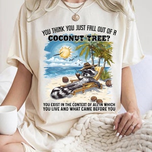 Puede incluir: Una camiseta blanca con un gráfico de un mapache sentado en una silla de playa con una bebida de coco. El texto en la camiseta dice "¿Crees que acabas de caer de un cocotero? Existes en el contexto de todo en lo que vives y lo que te precedió."