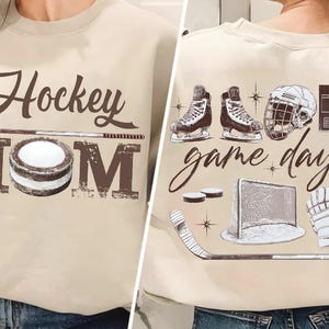 Puede incluir: Sudadera beige con texto marr&oacute;n y gr&aacute;ficos de hockey. La parte delantera presenta "Hockey Mom" con un palo y un disco de hockey. La parte trasera muestra patines, un casco y "game day" con equipamiento de hockey.