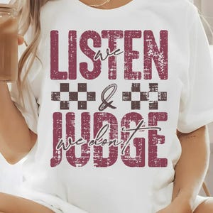 May include: White t-shirt with a maroon distressed graphic that says "Listen we don't judge" with a checkered pattern between the words.