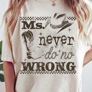 Peut inclure: T-shirt crème avec un motif graphique représentant un chapeau de cowboy, une botte de cowboy et le texte "Ms. never do no wrong" dans une police vieillie. Le motif comprend une bordure à carreaux.