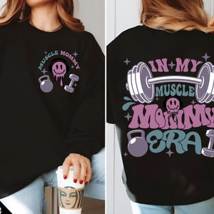 May include: Black sweatshirt with a pink and blue graphic design that says "Muscle Mommy Era" with a barbell, dumbbells, and a smiley face.