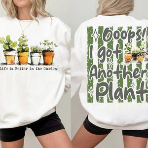 Könnte beinhalten: Weißes Sweatshirt mit einer Aquarellillustration von Topfpflanzen. Auf der Vorderseite des Sweatshirts steht der Text "Life is Better in the Garden". Auf der Rückseite des Sweatshirts steht der Text "Oops! I got Another Plant."