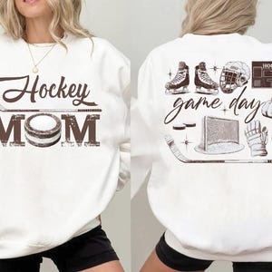 May include: White sweatshirt with a brown 'Hockey Mom' graphic on the front. The back features hockey-related illustrations and the words 'game day'. Includes images of skates, a helmet, a net, and gloves.